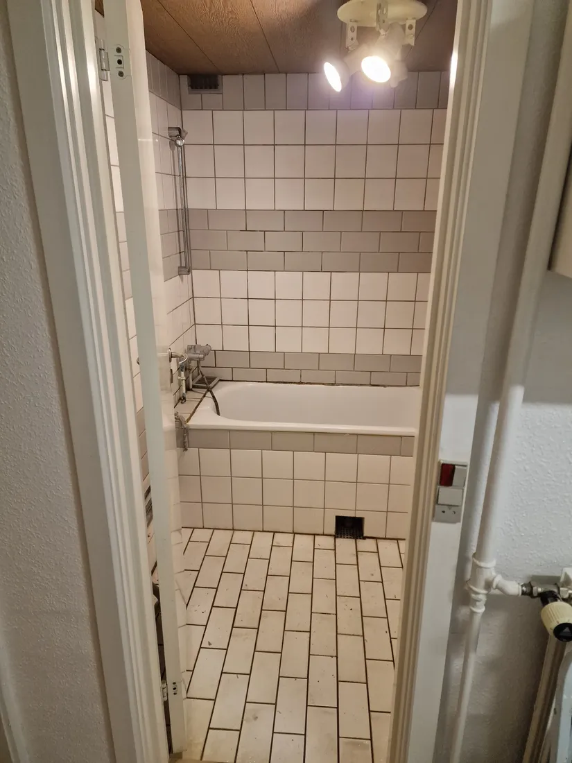 Gammelt slidt badeværelse med hvide fliser og badekar, før renovering